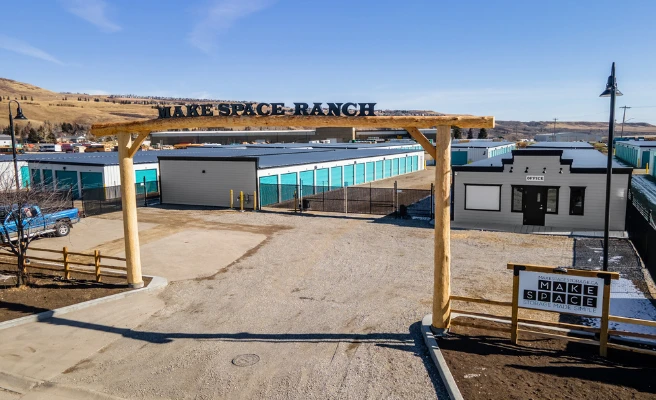 Entrance to a self-storage facility named ‘Make Space Ranch,’ featuring a large wooden archway with the name displayed on top, gravel driveway, and rows of teal-colored storage units in the background. A white office building and a sign reading ‘Make Space’ are visible under a clear blue sky with surrounding hills.